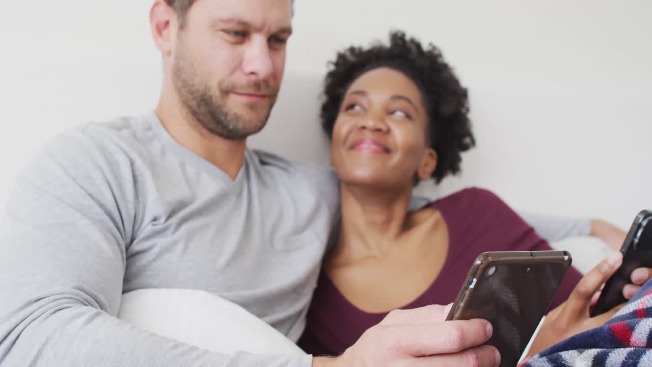 video de una feliz pareja diversa usando un teléfono inteligente en la cama y hablando