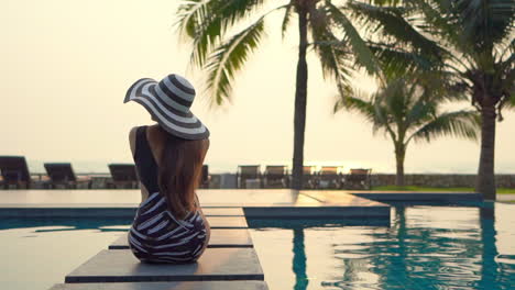 back view of a slim tourist lady sitting near a pool, wearing a black and white swiming suit and a floppy hat , admiring the view during sunset