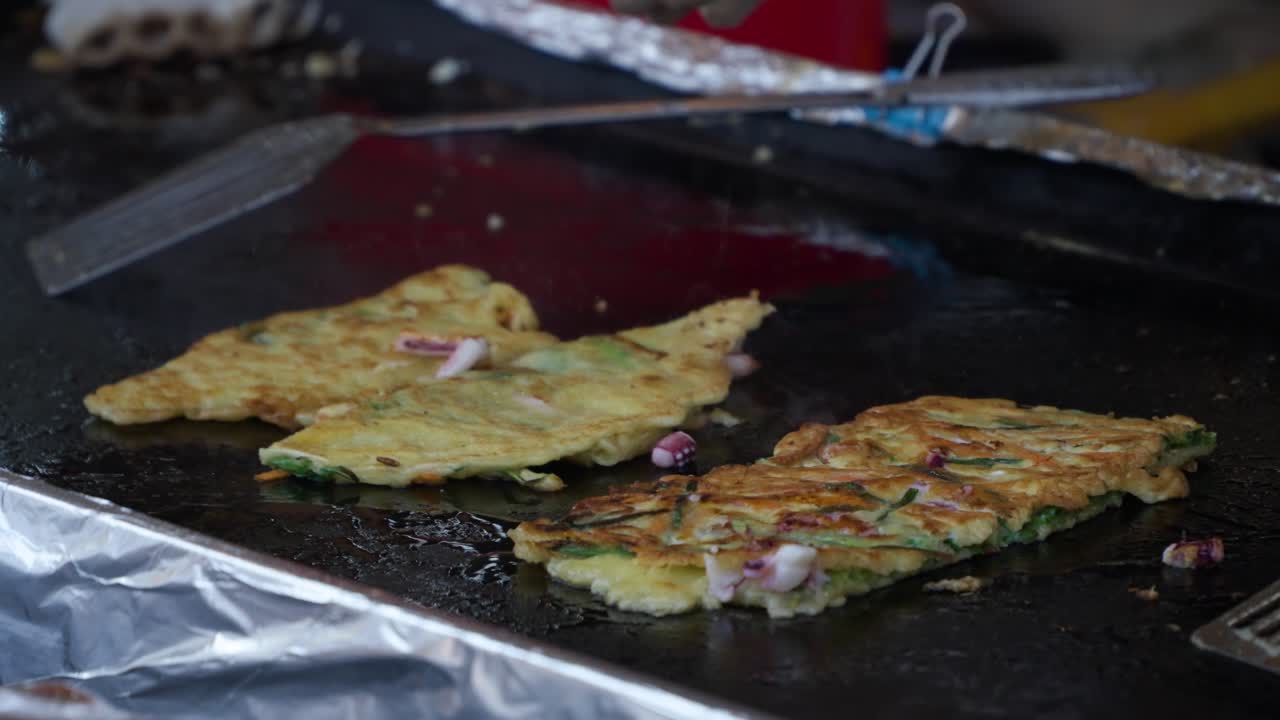 Close-up of a vendor's gloved hands using spatulas to flip and cook savory Korean street food pancakes on a large, shimmering metal grill at a local outdoor market