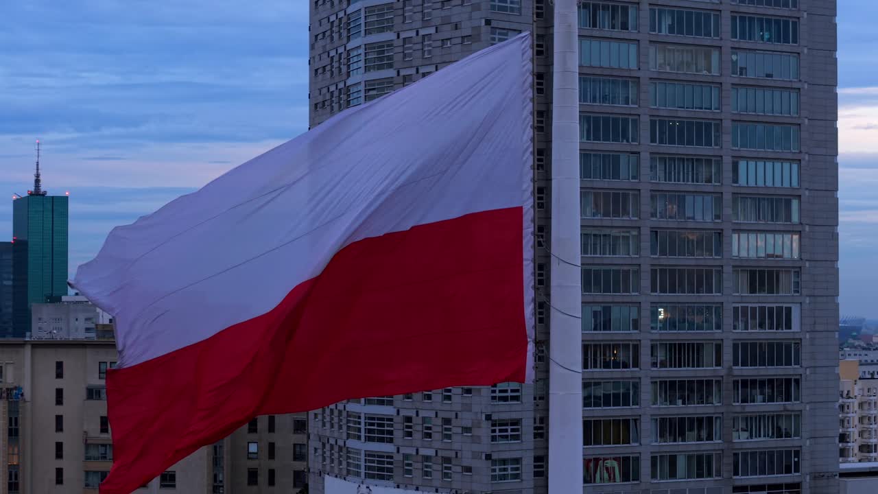 Slow motion aerial orbit of Polish flag waving in strong wind, Warsaw cityscape in the background