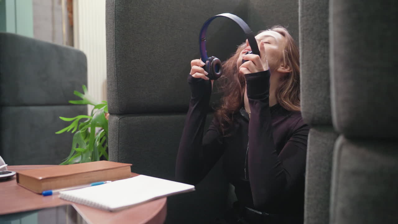 Lady with blond hair wearing headset gently closes book and begins removing headset while swinging head slightly in relaxed indoor setting with plants and soft lighting, creating a calm atmosphere