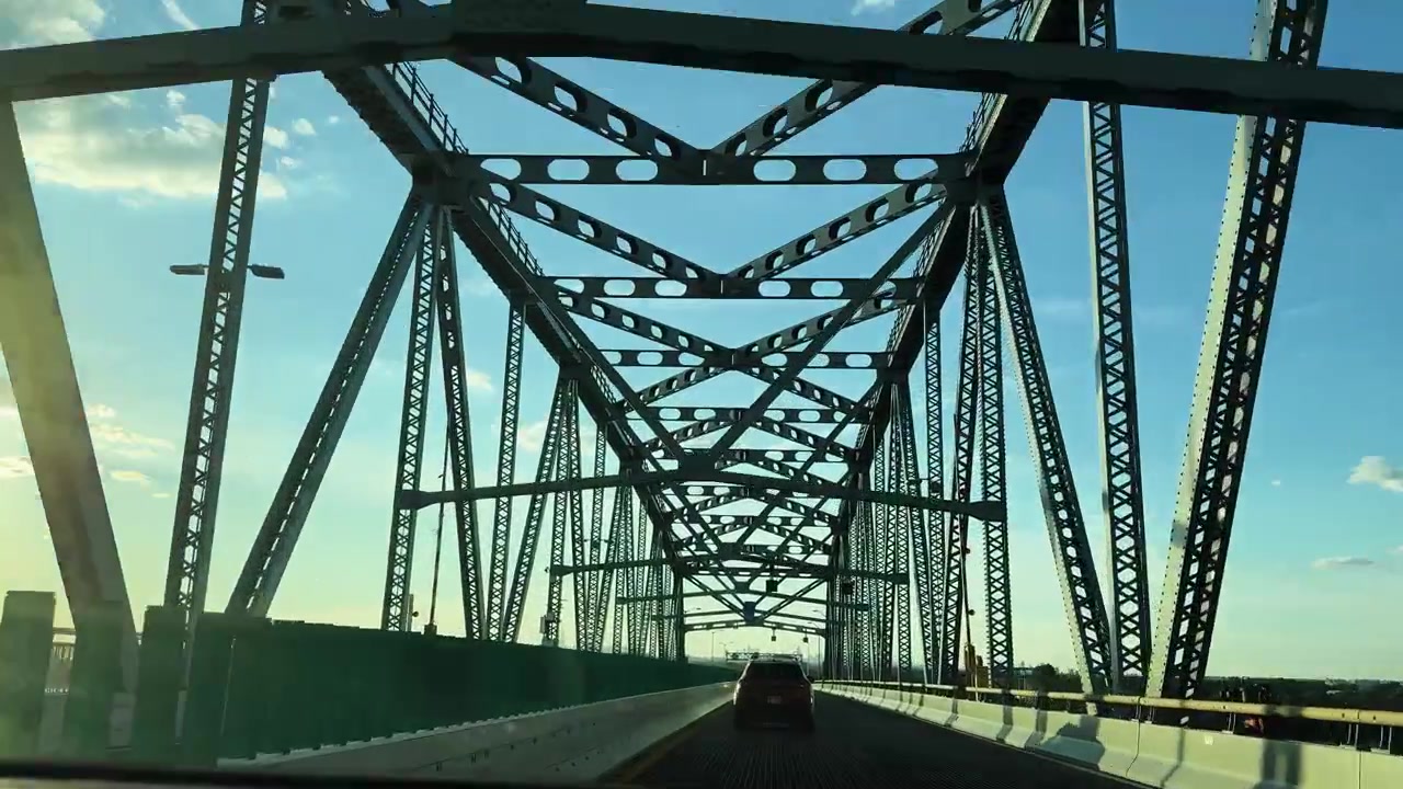 Vehicle drives across steel truss bridge at sunset with clear sky and clouds