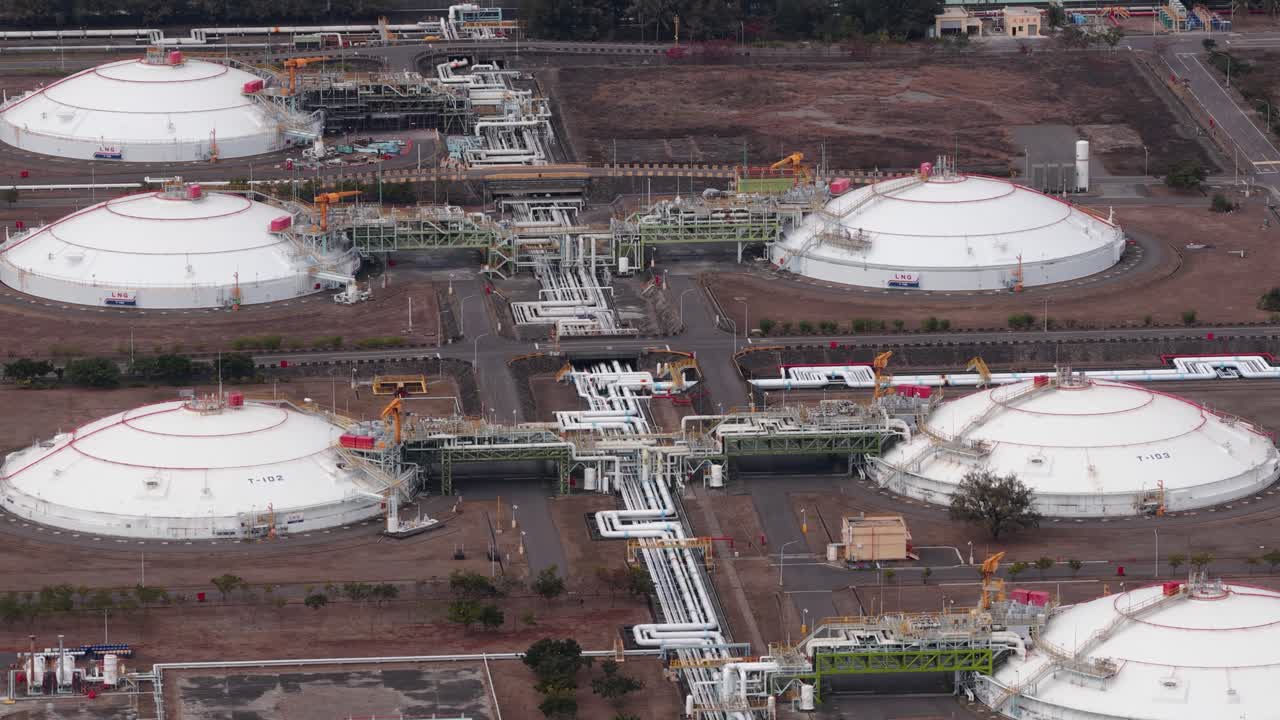 Büyük, beyaz depolama tankları, karmaşık boru hattı ağı ve enerji üretimi için çevresindeki endüstriyel altyapısı ile bir sıvılaştırılmış doğal gaz terminalini gösteren yüksek açılı bir perspektif.
