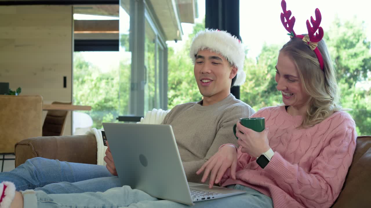 Coppia con cappello da Babbo Natale e cerchietto da renna che parla a bassa voce, digita sul portatile, sorseggia da una tazza e si appoggia al divano
