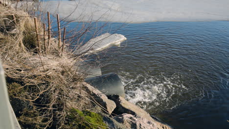 Slow-pan-of-water-flowing-from-sewage-pipes-into-icy-lake,-sunny-day