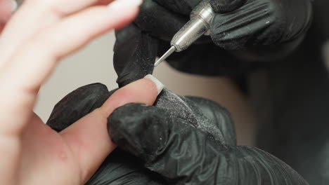 close up view of nail technician wearing black gloves using electric drill tool to gently file acrylic extension on client fingernail and dust particles visible