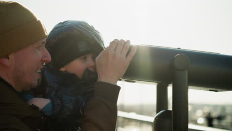 a father holds his son up to a telescope, guiding him as he peers through it with curiosity, the father smiles, supporting the telescope to ensure a steady view