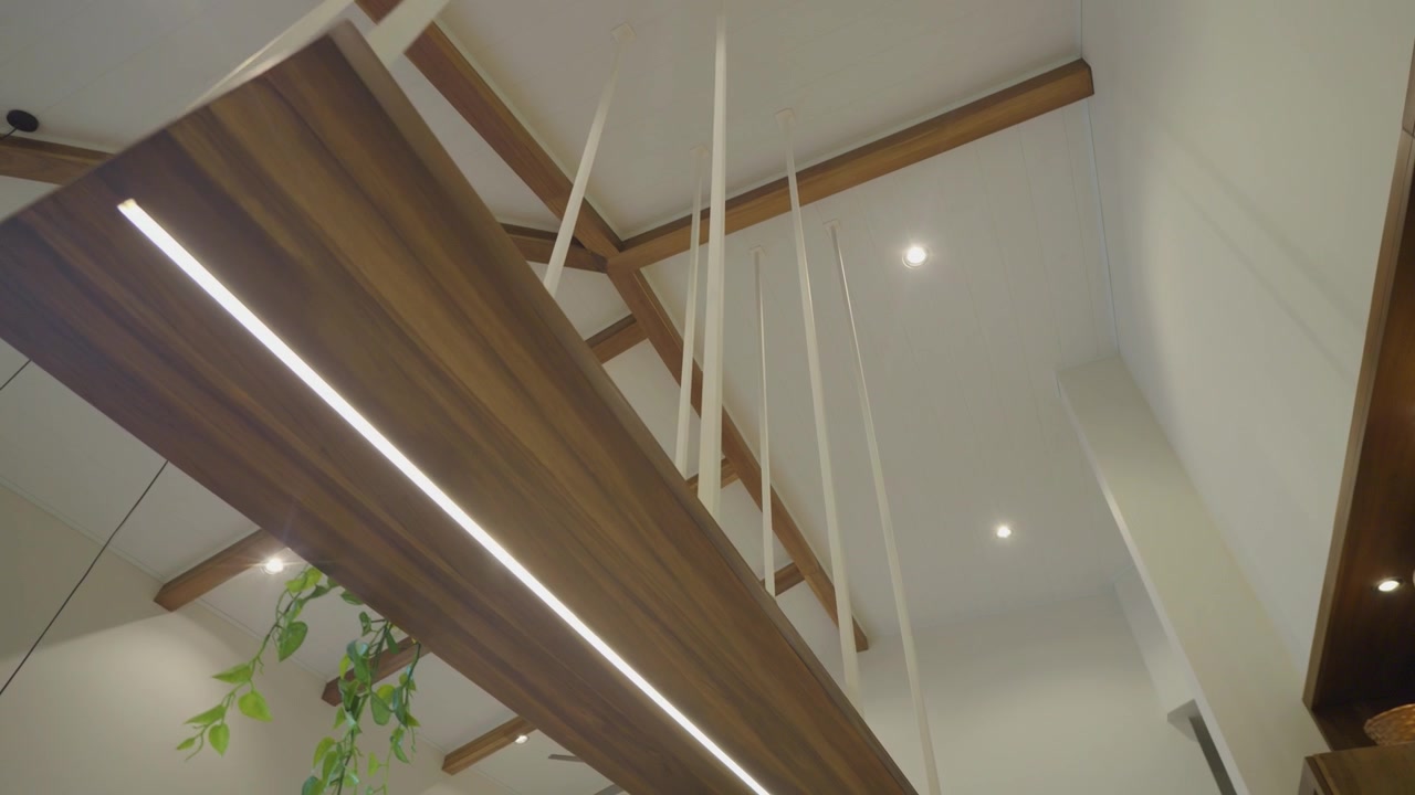 Modern room ceiling features white beams and a suspended wooden plank. Linear light strips run along the plank. Recessed ceiling lights illuminate the space. Green plant adds natural touch