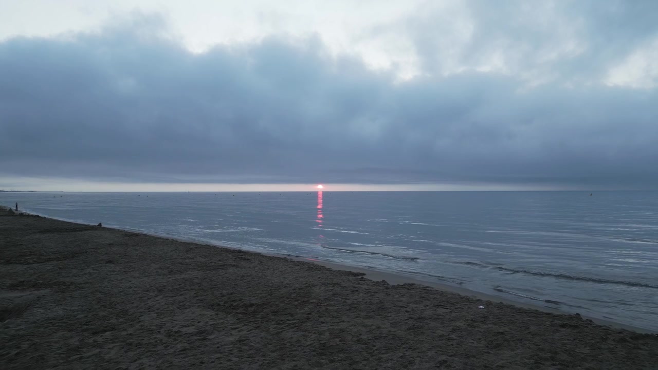 穏やかな海に赤い太陽が昇り、水面にその光を映し出している。曇り空が静謐な雰囲気を一層引き立て、人けのないビーチが孤独と安らぎの感覚を深めている。