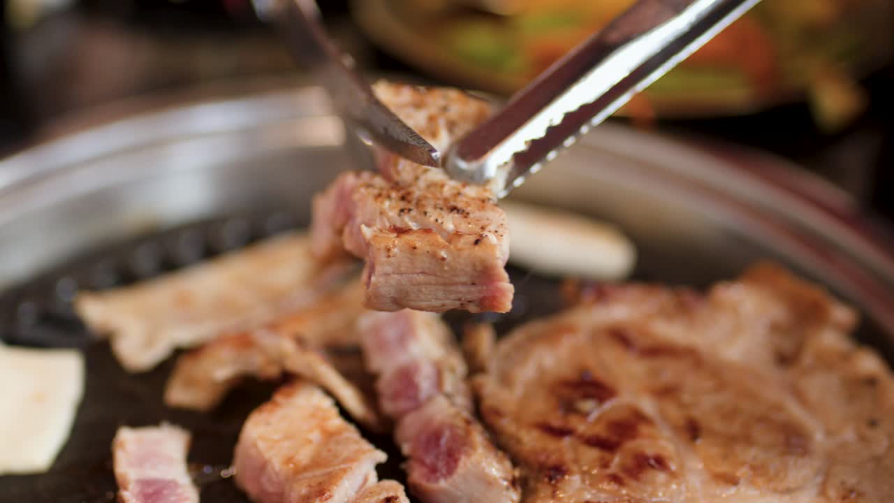 Hand uses scissors and tongs to cut grilled pork on tabletop Korean barbecue grill