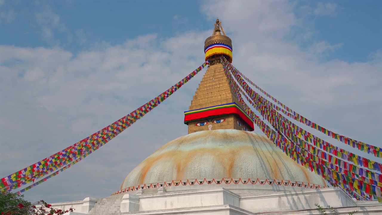 Staattinen laajakulmakuva, joka vangitsee ikonisen Boudhanathin stupan värikkäiden rukouslippujen levittäytyessä yli kauniin sinisen taivaan pehmeine valkoisine pilvineen Kathmandussa, Nepalissa.