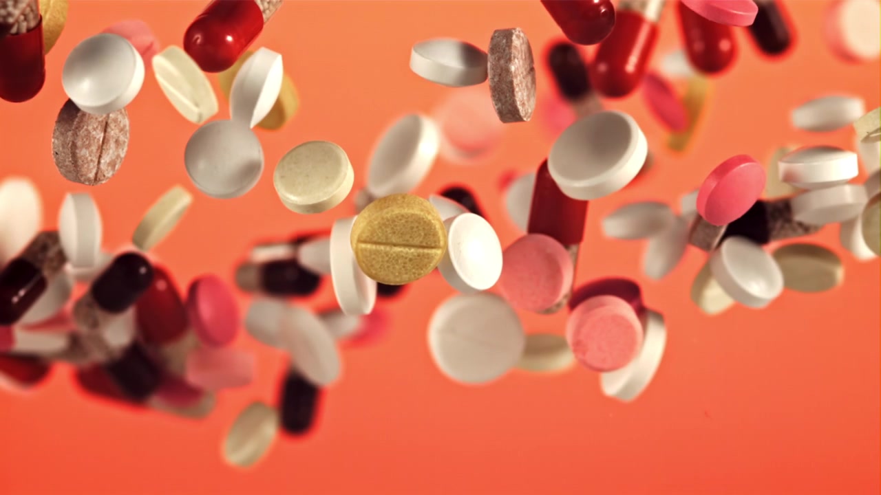 Pillole e capsule colorate che cadono al rallentatore su sfondo arancione.