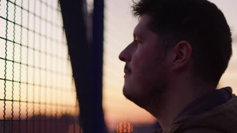 A sad man stares through a blue metal grate on an iron bridge, his face filled with contemplation. The dusk sky in the background enhances the emotional atmosphere of solitude and reflection.