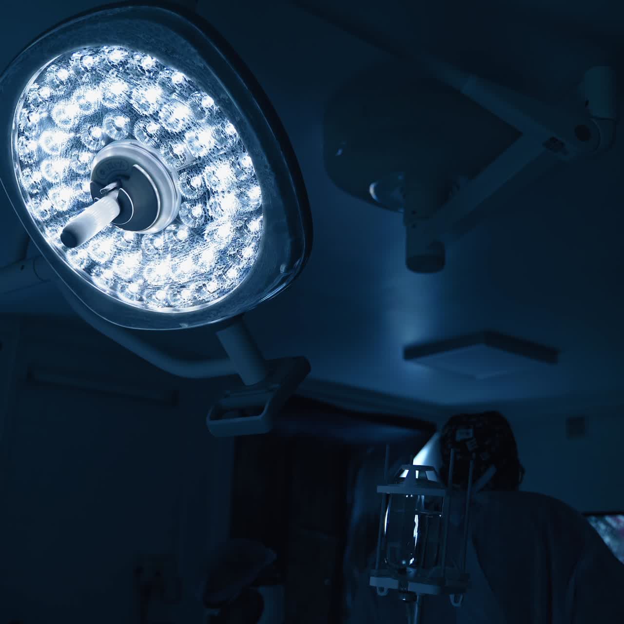 Surgical room in darkness. Medical lamp shines in the operating room. Doctor perform a surgery while looking on the screen of monitor.