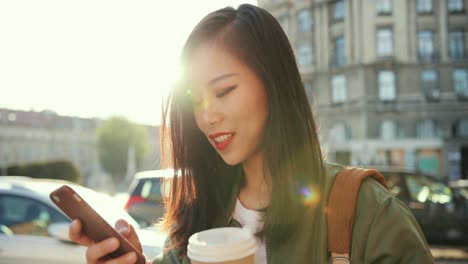 Cerca-De-La-Joven-Y-Bella-Mujer-Sonriente-Escribiendo-Un-Mensaje-En-El-Smartphone-Y-Riéndose-De-La-Ruidosa-Calle-De-La-Ciudad