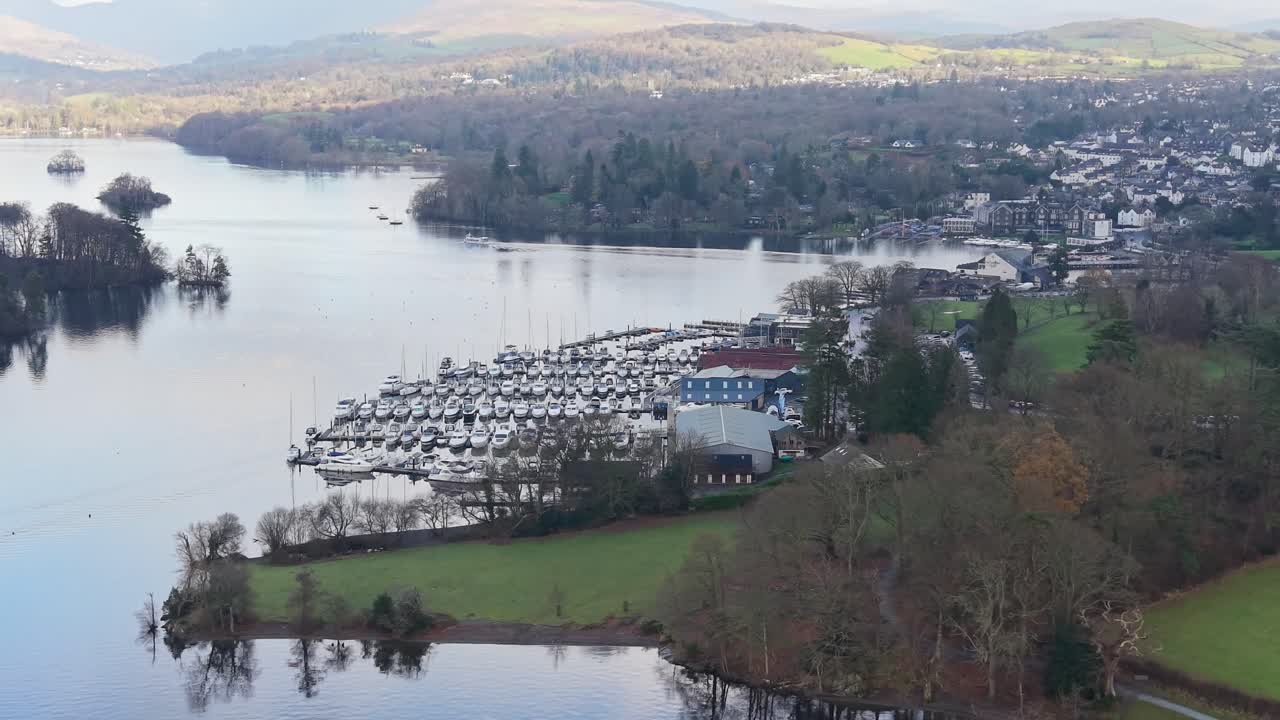 Drone melakukan pan ke kiri melewati marina di dekat pusat kota Bowness di Danau Windermere. Ponton paralel berada di samping garis pantai, dengan pepohonan musim dingin dan perbukitan yang bermandikan sinar matahari terlihat di kejauhan di bawah awan