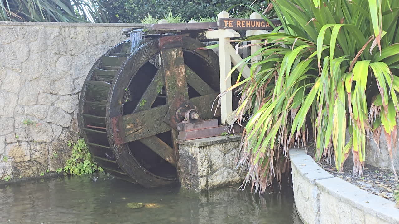 Static shot of old wooden waterwheel at historic mill with weathered stone wall and lush tropical plants with green foliage in background in Napier, New Zealand