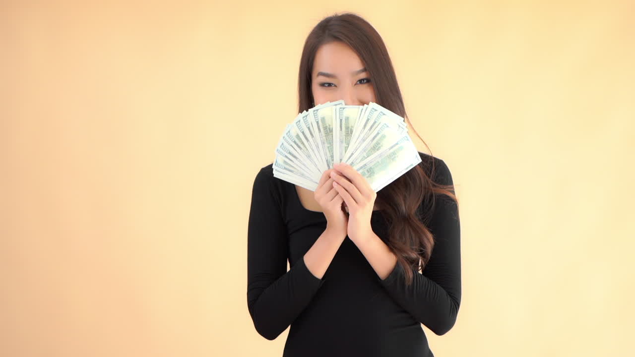 หญิงสาวชาวเอเชียผู้ร่ำรวยและมีความสุขซ่อนใบหน้าของเธอไว้หลังธนบัตรดอลลาร์ที่จัดเรียงเป็นรูปทรงพัด, การเคลื่อนไหวช้าแบบเต็มเฟรม