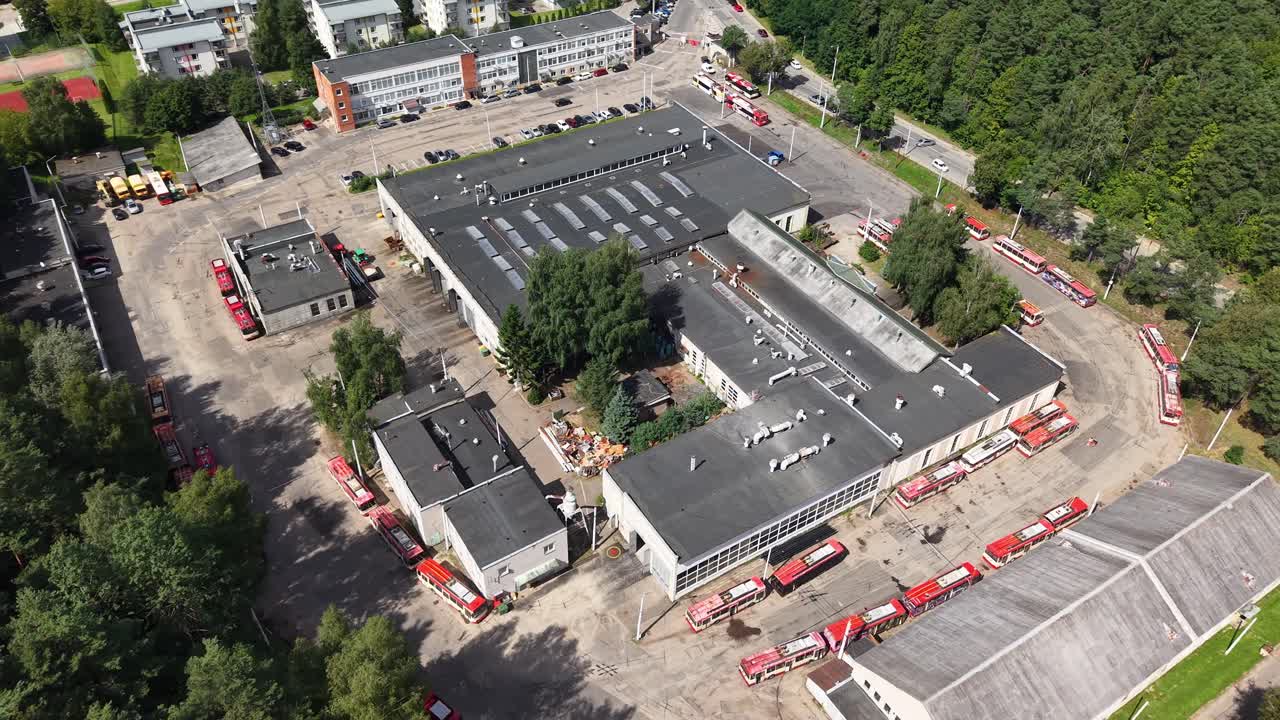 Wide drone establishing shot of the Vilnius trolleybus depot complex beside dense forest, showing maintenance buildings, paved yards, and red trolleybuses staged around the facility