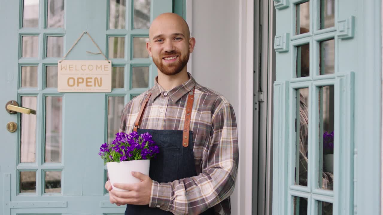 Concept of small business florist entrepreneur the charismatic man owner of the floral shop smiling large to the camera and holding a flowers pot in hands