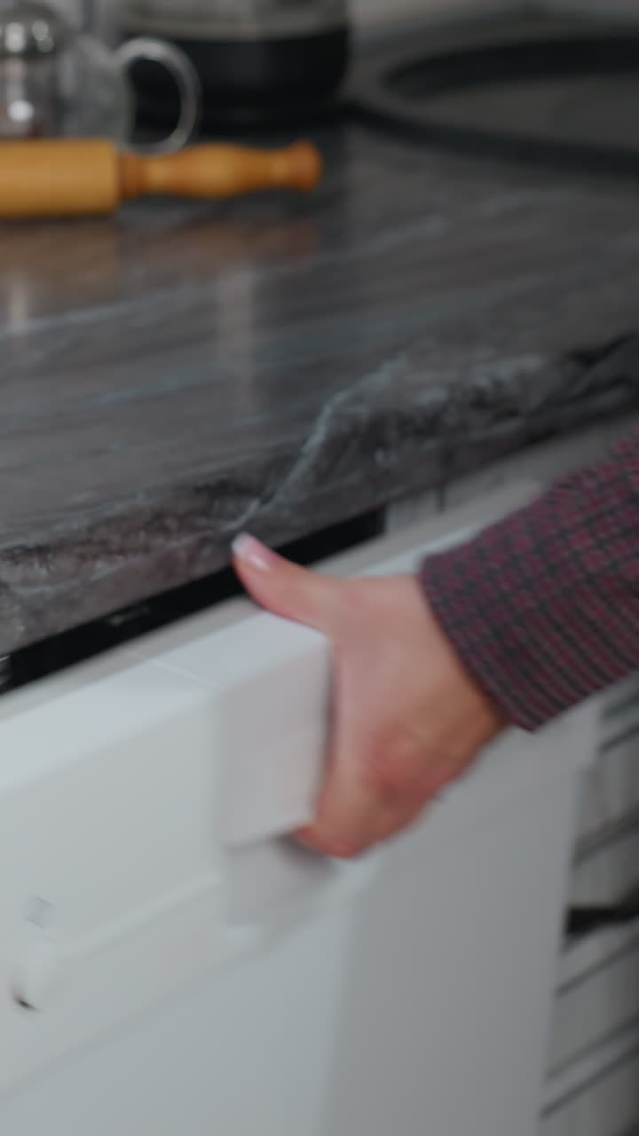Donna che prende una pentola da un mobile della cucina e la appoggia su un moderno piano cottura elettrico con display digitale, con oggetti sul piano di lavoro, lavastoviglie aperta, interni eleganti e ambiente domestico accogliente
