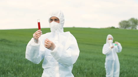 investigador caucásico con traje protector sosteniendo un tubo de ensayo mientras realiza el control de plagas en el campo verde