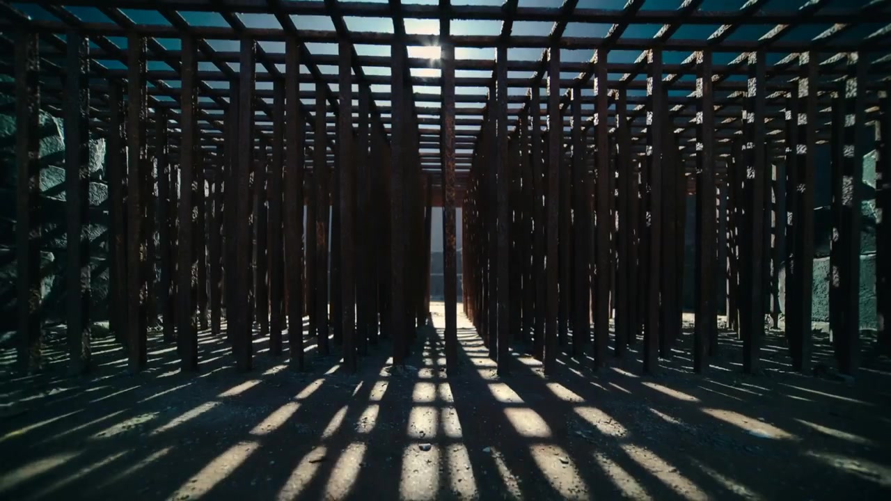Pali e travi che reagiscono al sole che entra da una fessura su un pavimento di terra all'aperto, con un bagliore dell'obiettivo che rivela l'uscita.