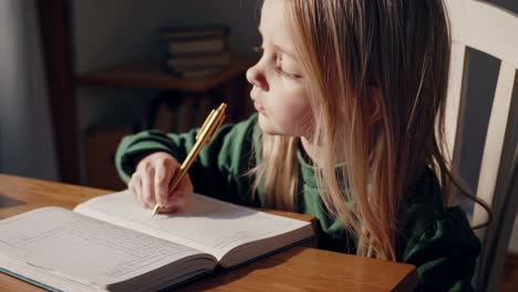 concentrated preschooler girl practices her writing skills, diligently filling pages of notebook with pen, sitting at wooden table in cozy home environment