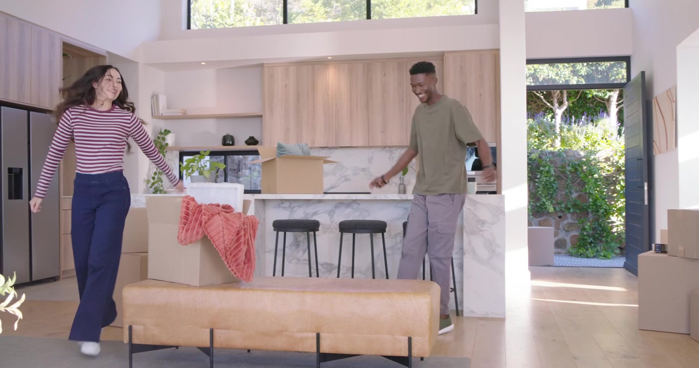 Couple unpacking at island, woman in stripes with phone spinning, man in tee dancing and embracing