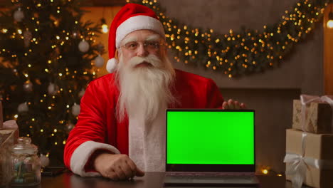 Der-Weihnachtsmann-Hält-Einen-Laptop-Mit-Einem-Grünen-Bildschirm,-Während-Er-In-Der-Weihnachtsdekoration-Sitzt.-Hochwertiges-4K-Filmmaterial