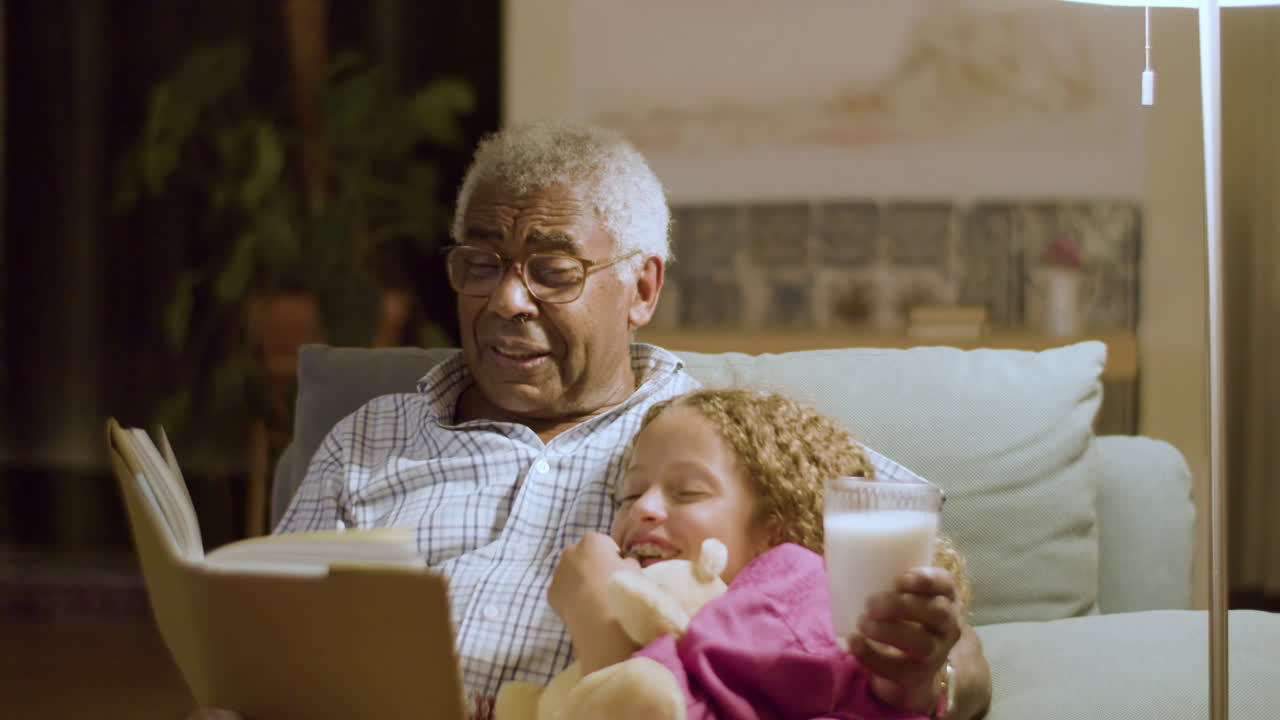 abuelo leyendo divertidas historias de buenas noches para su nieta mientras sostiene un vaso de leche