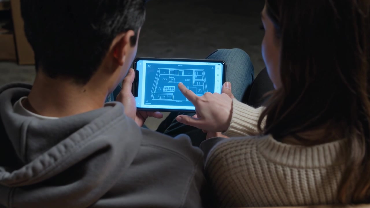 Coppia in felpa con cappuccio e maglione che guarda un progetto blu su tablet, indicando sul divano, tra le scatole, mentre pianificano un trasloco.