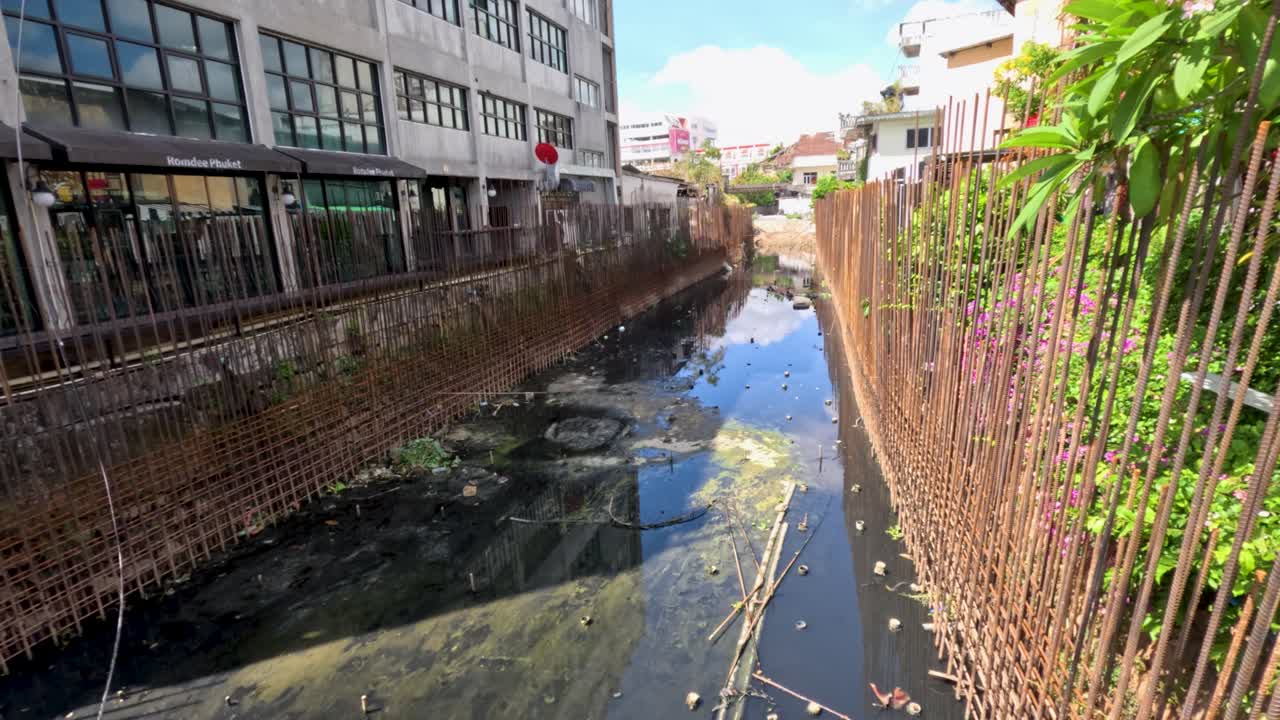 Daytime tracking shot of dirty water, debris, and rusty steel infrastructure in urban canal