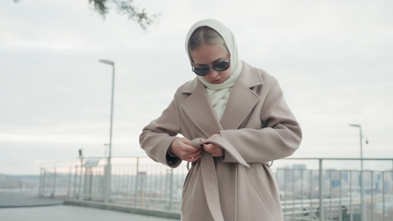 Dışarıda kıyafetini düzelten kadın. Açık havada kabanını sabitleyen ve vakur görünen modaya uygun kadın. Dışarıda kabanının kemerini bağlarken kendinden emin bir duruşla askısını kontrol eden şık kadın.