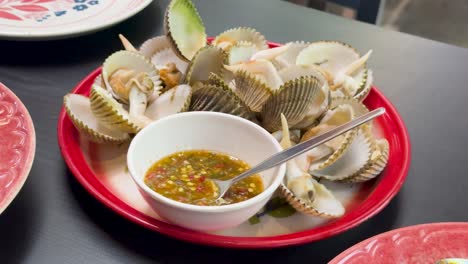 a red plate of steamed blood cockles served with a bowl of spicy thai seafood dipping sauce on a restaurant table under bright indoor lighting. subtle camera movement reveals the dish from multiple angles