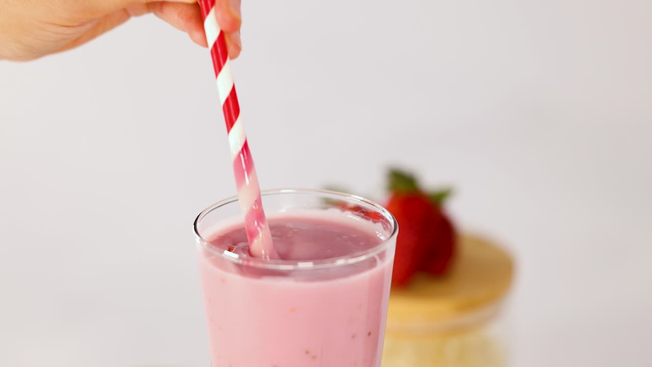 Hand stirs creamy strawberry banana smoothie in glass, bright lighting, close-up, minimal background