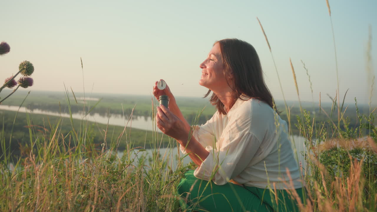 Sunset lit woman in green skirt squatting on grassy river bank blowing shimmering soap bubbles that drift above blurred valley with wild flower and thistle under warm golden light