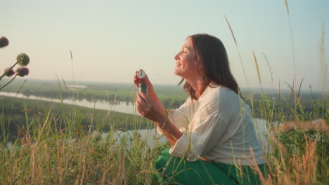 sunset lit woman in green skirt squatting on grassy river bank blowing shimmering soap bubbles that drift above blurred valley with wild flower and thistle under warm golden light