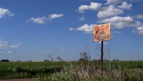 Se-Ve-Un-Cartel-Destrozado-Que-Advierte-De-Un-Canal-Profundo-En-Una-Zona-Rural-De-La-Provincia-De-Santa-Fe,-Argentina,-Con-Un-Campo-Sembrado-De-Soja-Al-Fondo