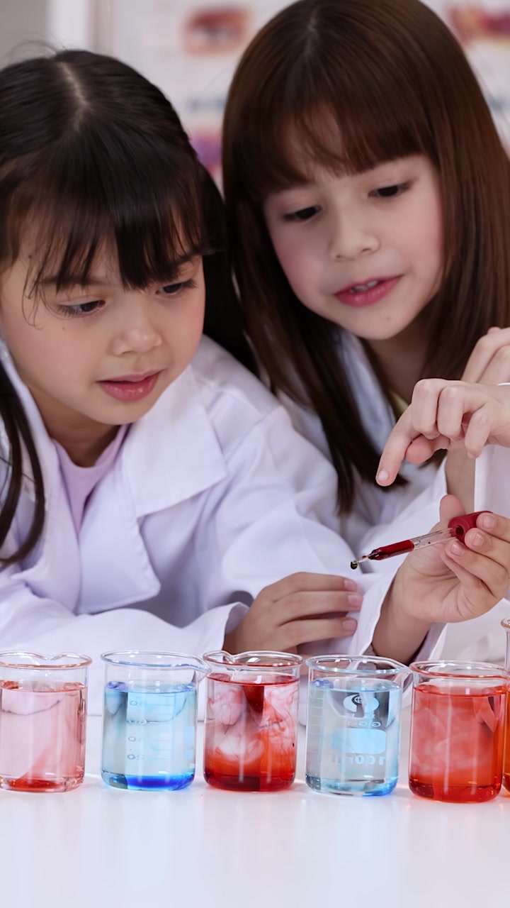 Two children in white lab coats use pipettes to drop liquid into colorful beakers.