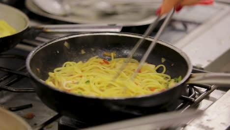 chef stirring spaghetti with vegetables in a pan over a gas burner