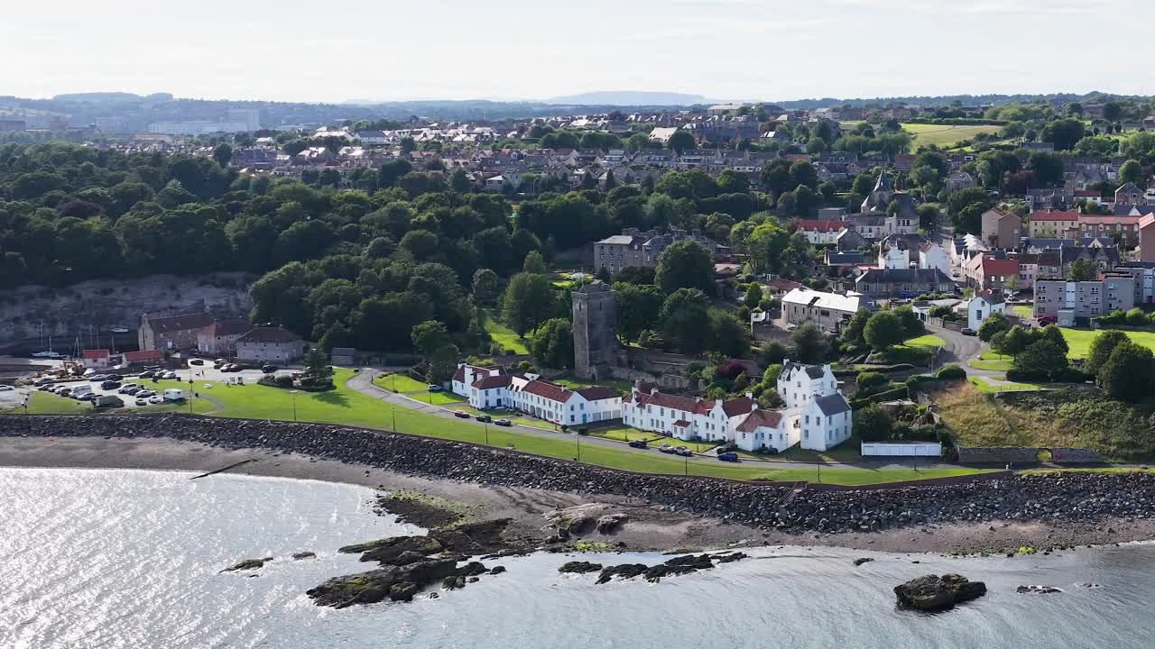 Drone camera smoothly pans across seaside town, historic buildings, shoreline, and lush green landscape