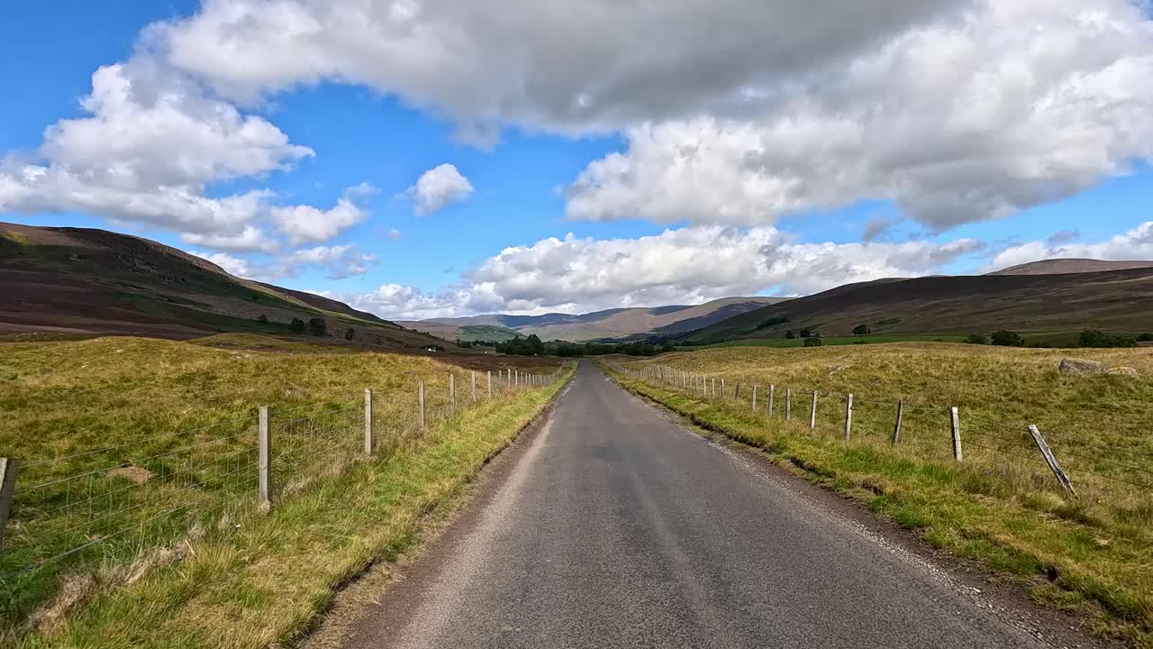 A vehicle travels down a winding country road through scenic Scottish hills under partly cloudy skies, with dynamic forward camera movement and natural daylight