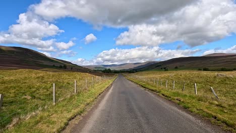 a vehicle travels down a winding country road through scenic scottish hills under partly cloudy skies, with dynamic forward camera movement and natural daylight