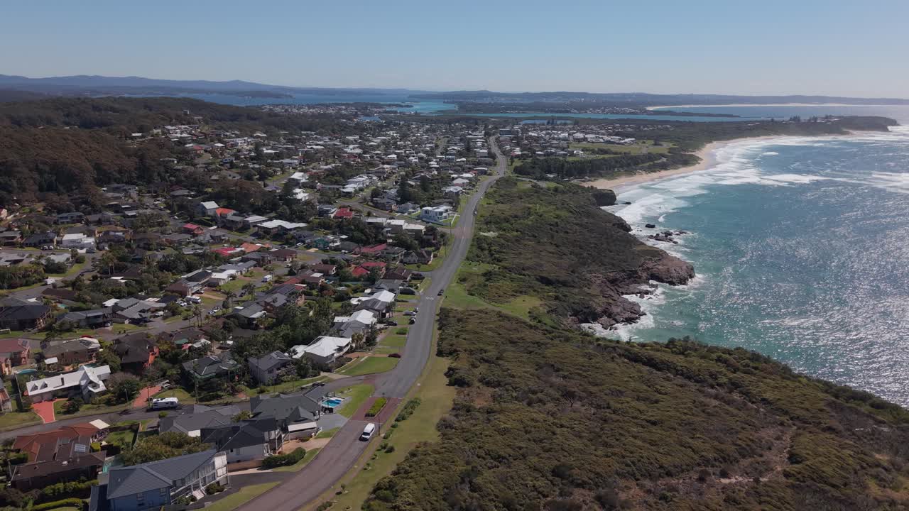 Drone establishing over road above open sea over rooftops and beach, revealing tranquil water and pastel sky, NSW Australia