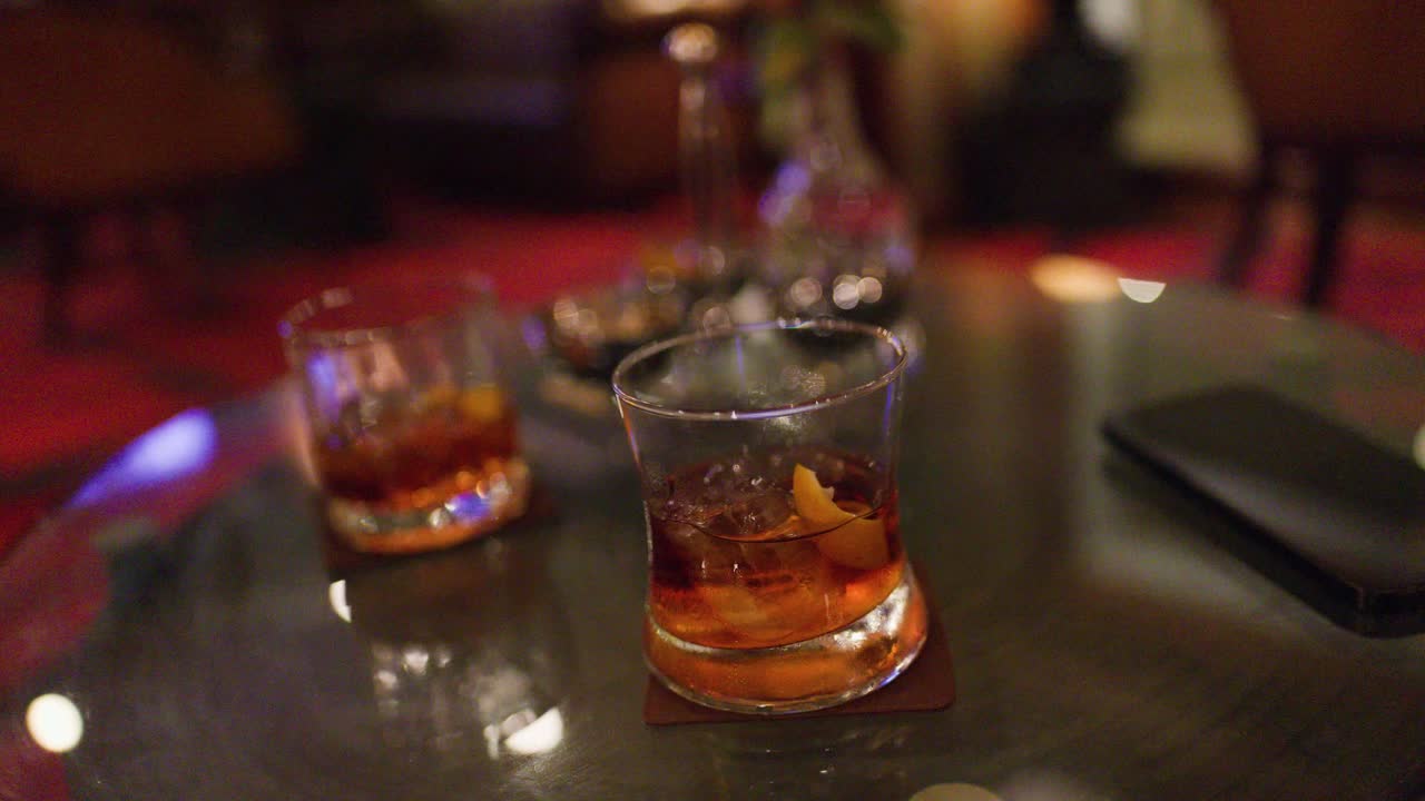 Two whiskey cocktails with ice on glass table in dimly lit, elegant bar lounge environment