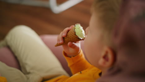 close up side view of little boy in orange shirt relaxing barefoot on pink beanbag while holding ice cream cone, enjoying sweet treat in cozy indoor setting