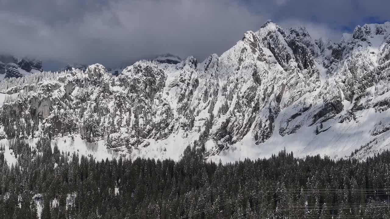 Drone clip showing snow covered mountain range with alpine forest below