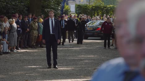 security guard stands watch during memorial event at oosterbeek war cemetery, surrounded by soldiers and attendees paying tribute. solemn, with people walking in the background. oosterbeek, gelderland