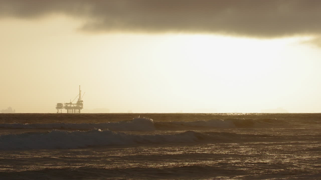 Bølger, der slår ind i slowmotion med en olieboreplatform i baggrunden ved solnedgang